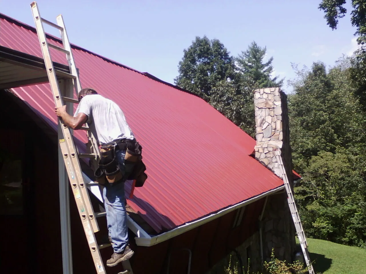 Roofer on a ladder beside a red metal roof and stone chimney — Soffit & Fascia Repair by Roofing Brothers in Bowlus